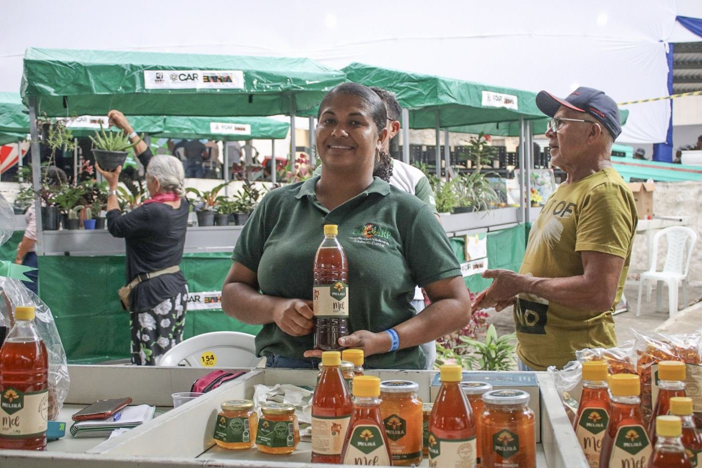 Associada sorrindo atrás da barraca com mel e doces na Feira Municipal de Galileia